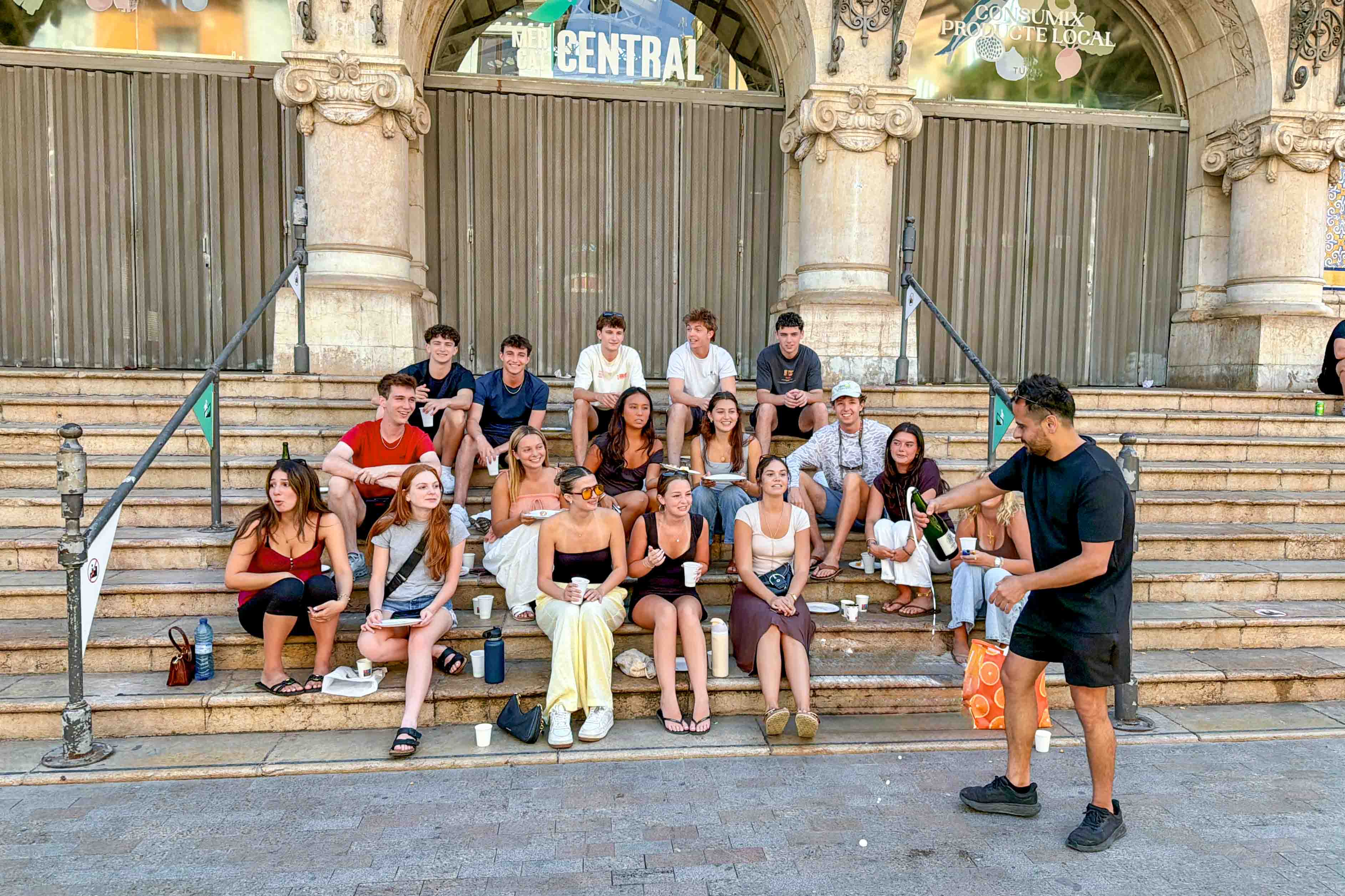 A faculty member opening a bottle of champagne in front of his students on the steps laughing.